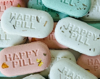 May include: Close-up of several pastel-colored bath bombs. Each oval-shaped bomb is imprinted with the words "HAPPY PILL" and decorated with dried botanicals. The bath bombs are in shades of pink, blue, and white.