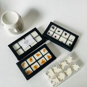 May include: An assortment of natural soy wax melts in black boxes and a clear plastic container. Some melts have dried flowers or fruit pieces. A white ceramic oil warmer is in the upper left corner. The text "Natural Soy Wax Melts" is visible.