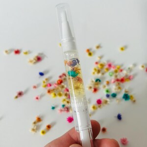 May include: A clear, white pen-shaped nail cuticle oil applicator filled with colorful dried flowers. The applicator has a clear cap and a white brush tip. Scattered around the applicator are more dried flowers in various colors.