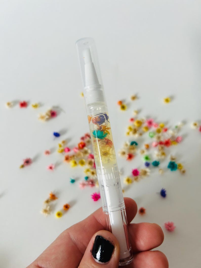 May include: A clear, white pen-shaped nail cuticle oil applicator filled with colorful dried flowers. The applicator has a clear cap and a white brush tip. Scattered around the applicator are more dried flowers in various colors.