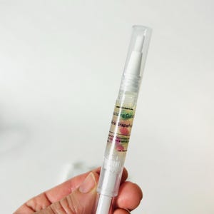 May include: A clear cuticle oil pen with a white applicator tip and a clear cap. The pen has a label with the text "Botanical" and "Pink Grapefruit". The oil contains pink grapefruit pieces.