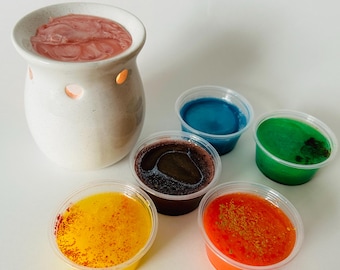 May include: A white ceramic essential oil warmer with a pink wax melt, alongside six small plastic containers filled with colorful wax melts in yellow, orange, brown, blue, and green. The warmer has three small openings.