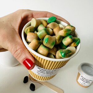 May include: A white paper cup filled with green and brown soy wax melts shaped like coffee beans. The cup has a brown paper sleeve with the text "All Natural Soy Wax Melts Coffee Beans" printed on it.