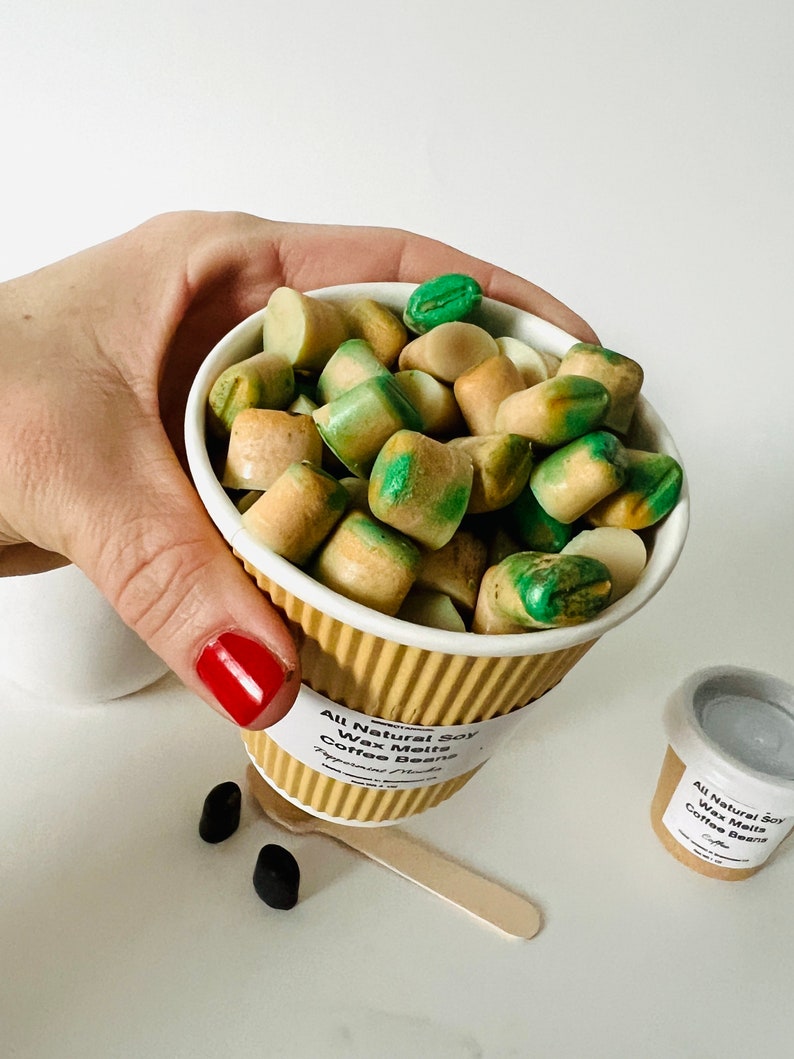 May include: A white paper cup filled with green and brown soy wax melts shaped like coffee beans. The cup has a brown paper sleeve with the text "All Natural Soy Wax Melts Coffee Beans" printed on it.