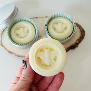 May include: Three round, light yellow solid lotion bars with a bee design, one held in a hand. Two bars are in small, silver tins with a corrugated paper liner. The bars are on a wooden surface.