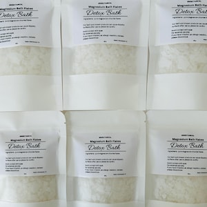 May include: Six white stand-up pouches containing Magnesium Bath Flakes Detox Bath. Each label lists ingredients as pure magnesium chloride flakes, safety warnings, and states 200g. The pouches have a clear window showing white flakes.