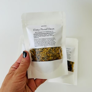 May include: A white pouch with a clear window showing a blend of dried herbs and flowers. The pouch is labeled "Detox Facial Steam with Peppermint Essential Oil". The ingredients are listed as chamomile flowers, calendula flowers, lavender buds, rosemary flowers, lemongrass, peppermint flowers, fenugreek leaves, and peppermint essential oil. The directions are to place 2-3 teaspoons of the blend in a bowl with 3 cups of boiling water, cover and let steam for 5 minutes. Place a towel over your head, with eyes closed, lean over the steaming herbs about 10 inches from the edge of the pot, close enough to feel the steam but not close enough to be burned. Relax deeply for 10 minutes. Follow with a cool water rinse. 0.5 oz.