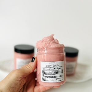 May include: A jar of pink body scrub labeled "Warm Vanilla Sugar" is held in front of a white background. The scrub is in a clear jar with a black lid. The label includes the brand name "MWBotanical". Two other jars are visible in the background.