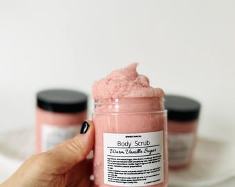 May include: A pink body scrub in a clear jar with a white label that reads "Body Scrub Warm Vanilla Sugar" and lists ingredients. The jar is being held by a hand.
