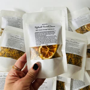 May include: A white pouch with a clear window showing dried orange slices and yellow herbs. The pouch is labeled "Refresh Facial Steam with Sweet Orange Essential Oil". The ingredients are listed on the back of the pouch.