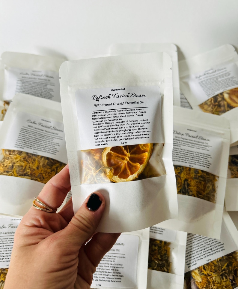 May include: A white pouch with a clear window showing dried orange slices and yellow herbs. The pouch is labeled "Refresh Facial Steam with Sweet Orange Essential Oil". The ingredients are listed on the back of the pouch.