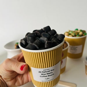May include: A brown paper cup filled with black soy wax melts shaped like coffee beans. The cup has a white label that reads "All Natural Soy Wax Melts Coffee Beans".