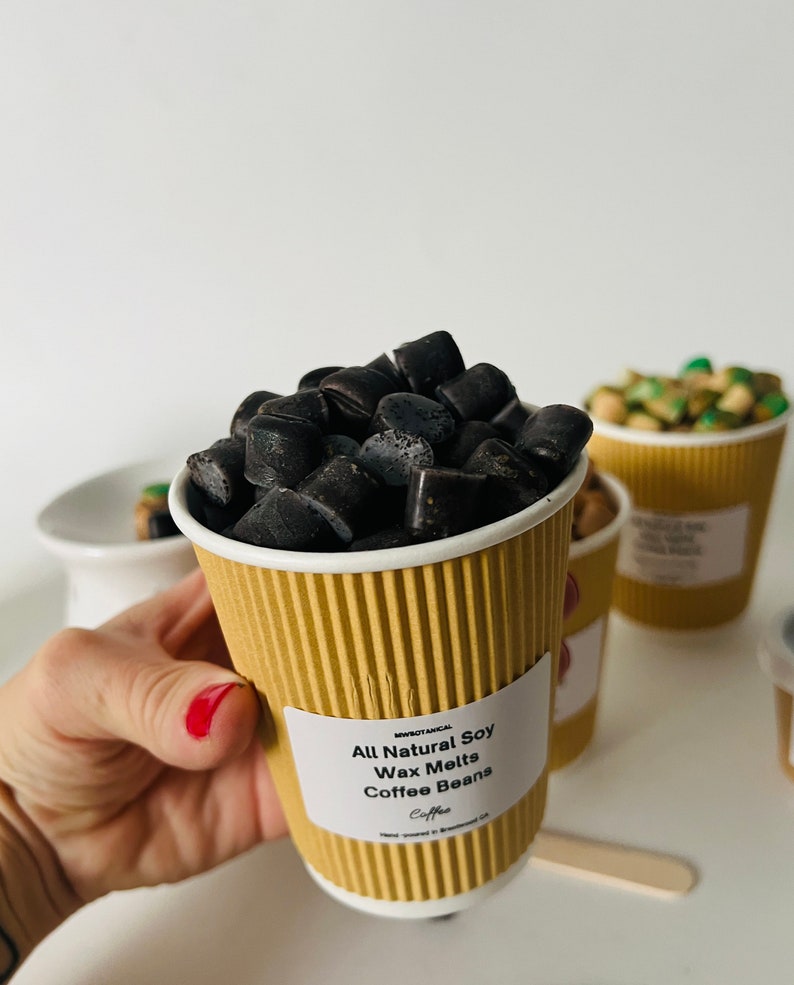 May include: A brown paper cup filled with black soy wax melts shaped like coffee beans. The cup has a white label that reads "All Natural Soy Wax Melts Coffee Beans".