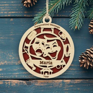 May include: A circular wooden ornament featuring a cutout design of theater masks, a ticket, and a stage. The center banner displays the name Maria, with the text Personalized Message at the top and Year at the bottom, set against a dark red background.