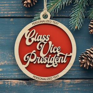 May include: A circular wooden Christmas ornament featuring a red center with the laser-cut text Class Vice President in a script font. The ornament has a light wood border with the words PERSONALIZED TEXT at the top and bottom and a twine loop.