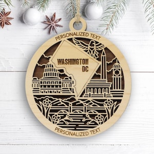 May include: A circular wooden ornament featuring a laser-cut design of Washington DC landmarks, including the Capitol building and Washington Monument. The text reads PERSONALIZED TEXT, WASHINGTON DC, and PERSONALIZED TEXT on a light wood finish.