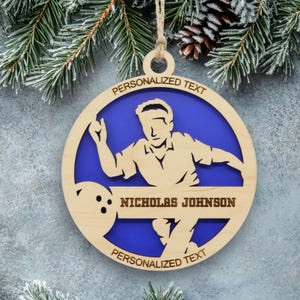 May include: A circular wooden ornament featuring a cutout silhouette of a person bowling with a ball. The design includes a blue background, a horizontal bar with the text NICHOLAS JOHNSON, and space for personalized text at the top and bottom.