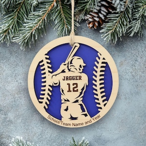 May include: A round wooden ornament featuring a cutout of a baseball player holding a bat, with the name JAGGER and number 12 engraved on the jersey. The design includes baseball stitching patterns and the text School/Team Name and Year.