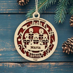 May include: A circular wooden Christmas ornament featuring a laser-cut choir of five people singing with musical notes and symbols. The center banner displays the name Maria, with text at the top reading Personalized Message and Year at the bottom.
