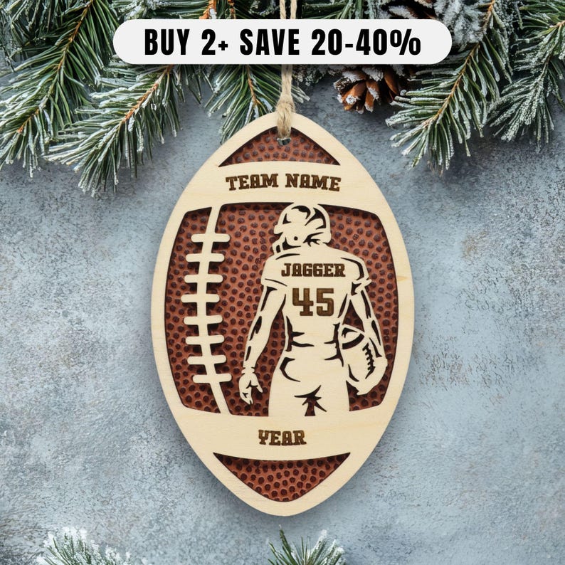 May include: A wooden football-shaped ornament with a cutout design featuring a football player. The ornament has the text "TEAM NAME" at the top and "YEAR" at the bottom. The player's jersey displays "JAGGER" and the number "45".