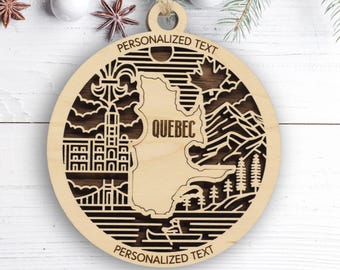 May include: A circular wooden ornament featuring a laser-cut design of the Quebec province map, a building, mountains, trees, and a canoe. The text "PERSONALIZED TEXT" appears at the top and bottom, with "QUEBEC" centered on the map cutout.