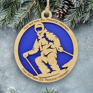 May include: A circular wooden ornament featuring a cutout of a skier in motion against a solid blue background. The wooden frame includes the text Your text here (Ex: Personal message) at the top and bottom, with a jute string loop for hanging.