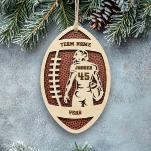 May include: An oval-shaped wooden ornament featuring a football design with a textured brown background. The cutout displays a football player silhouette with the text "TEAM NAME", "JAGGER", "45", and "YEAR" engraved on the light wood surface.