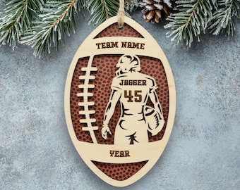 May include: An oval-shaped wooden ornament featuring a football design with a textured brown background. The cutout displays a football player silhouette with the text "TEAM NAME", "JAGGER", "45", and "YEAR" engraved on the light wood surface.