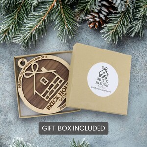 a wooden ornament with a house design, placed inside a gift box with a label that includes the text "GIFT BOX INCLUDED".