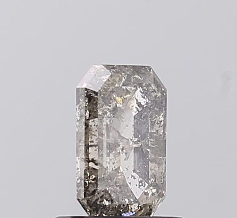 May include: A rectangular-cut gemstone with a grey and clear appearance. The gem has a textured surface and multiple facets, reflecting light. The gemstone is set against a plain, light background.
