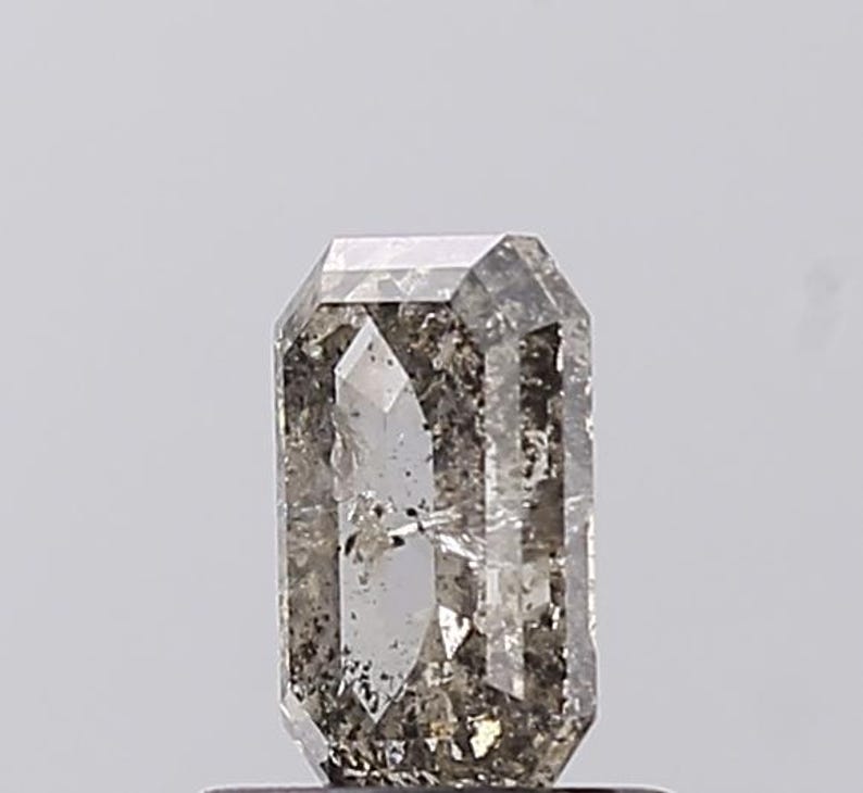 May include: A rectangular-cut gemstone with a clear, grey colour and internal inclusions. The gem has a polished surface and a symmetrical cut, reflecting light. The overall appearance is a mix of transparency and subtle colour variations.