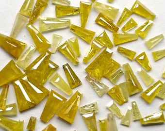 May include: A collection of yellow, faceted, rough cut, diamond shaped gemstones. The stones are various sizes and shapes, and are scattered on a white background.