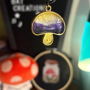 Puede incluir: Colgante de hongo dorado con cordón negro. La tapa del hongo está rellena de resina amarilla, blanca y morada. El tallo del hongo tiene una cara sonriente. El fondo incluye un juguete de peluche de hongo y un bordado enmarcado.