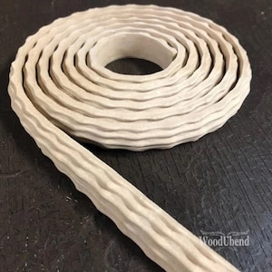 Könnte beinhalten: Eine aufgerollte, beige strukturierte Holzleiste mit einem wellenförmigen Muster. Ein Teil der Leiste ist abgerollt und erstreckt sich nach links unten. Das WoodUbend-Logo ist unten rechts sichtbar.