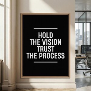 May include: A rectangular framed wall art piece featuring a black background with white text that reads HOLD THE VISION TRUST THE PROCESS, centered between two horizontal white lines, enclosed in a light wood frame.
