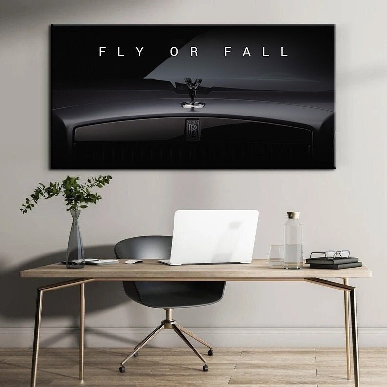 Puede incluir: Una impresi&oacute;n en lienzo en blanco y negro con el texto "FLY OR FALL" sobre un primer plano del cap&oacute; de un coche de lujo. El emblema y el adorno del cap&oacute; del coche son visibles. La impresi&oacute;n est&aacute; sobre un escritorio.
