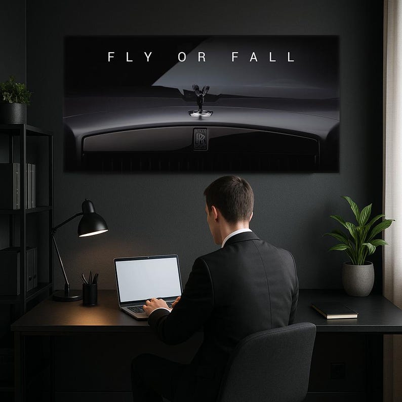 Puede incluir: Una impresi&oacute;n en lienzo en blanco y negro con el texto "FLY OR FALL" sobre un adorno de cap&oacute; Rolls-Royce. Una persona con un traje negro est&aacute; sentada en un escritorio con una computadora port&aacute;til. Tambi&eacute;n se ven una l&aacute;mpara de escritorio negra y una planta en maceta.
