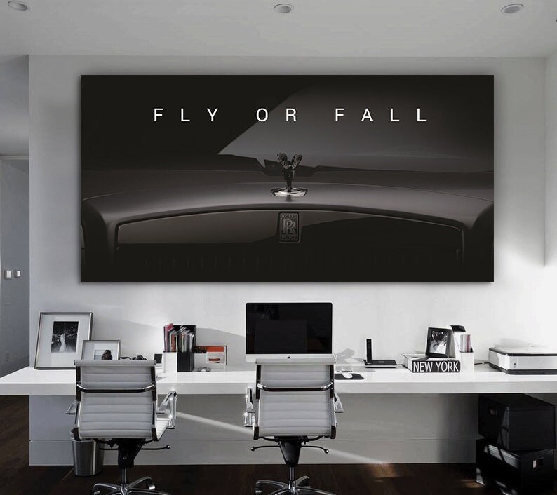 Puede incluir: Un lienzo en blanco y negro con un adorno de cap&oacute; de Rolls-Royce y el texto "FLY OR FALL". La imagen est&aacute; sobre un escritorio blanco con dos sillas de oficina, un ordenador y material de oficina. El tema es el lujo y la motivaci&oacute;n.