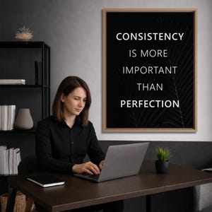 K&ouml;nnte beinhalten: Eine Frau in einem schwarzen Hemd benutzt einen Laptop an einem dunkelbraunen Schreibtisch. Ein gerahmter schwarzer Druck mit wei&szlig;em Text "CONSISTENCY IS MORE IMPORTANT THAN PERFECTION" h&auml;ngt an der Wand. Eine kleine Topfpflanze steht auf dem Schreibtisch.