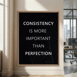 K&ouml;nnte beinhalten: Ein gerahmter Kunstdruck mit dem Text "CONSISTENCY IS MORE IMPORTANT THAN PERFECTION" in Wei&szlig; auf schwarzem Hintergrund. Der Druck hat einen hellbraunen Holzrahmen und ein subtiles Blattdesign. Das Kunstwerk wird in einem gut beleuchteten Raum ausgestellt.