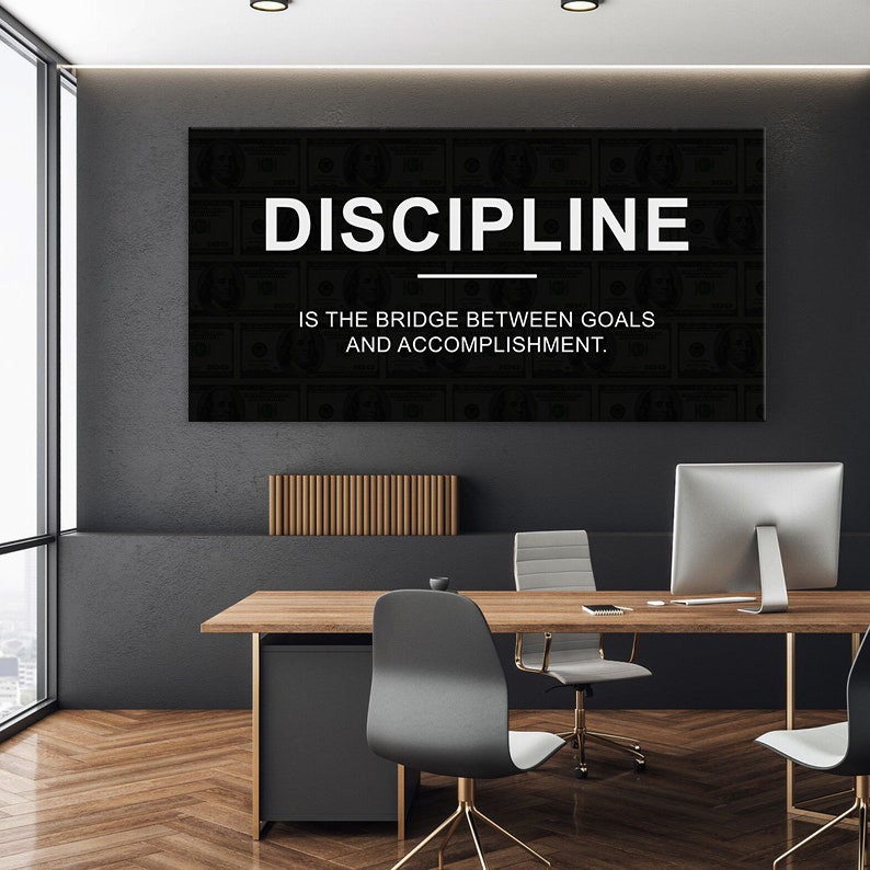 May include: A black and white motivational poster with the text "DISCIPLINE IS THE BRIDGE BETWEEN GOALS AND ACCOMPLISHMENT." The poster is hanging on a black wall in an office setting.