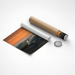 May include: A poster print featuring a car on a road with the text "ONE DAY REALITY WINS" in white. The print is partially rolled up with a white paper and a brown cardboard tube. A small, silver, round object is also present.