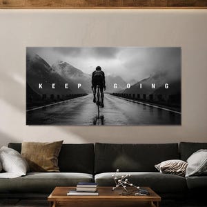 Könnte beinhalten: Ein rechteckiger Wandkunstdruck in Schwarz-Weiß, der einen Radfahrer zeigt, der auf einer nassen Straße in Richtung nebliger Berge fährt. Der Text KEEP GOING ist in weißen Großbuchstaben in der Mitte der Landschaft abgebildet.