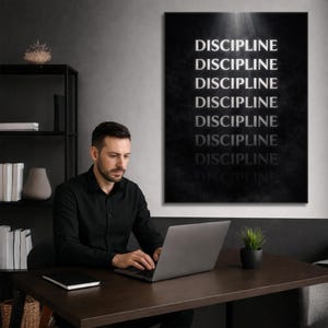 May include: A black and white wall art print with the word "DISCIPLINE" repeated vertically. A man in a black shirt is seated at a dark brown desk, working on a laptop. A small potted plant sits on the desk.