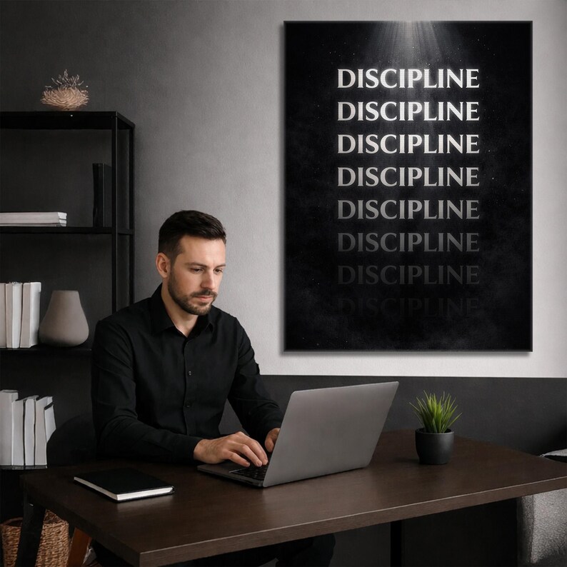 May include: A black and white wall art print with the word "DISCIPLINE" repeated vertically. A man in a black shirt is seated at a dark brown desk, working on a laptop. A small potted plant sits on the desk.