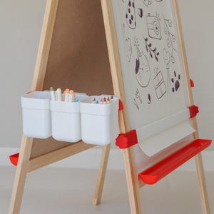 May include: A wooden art easel with a roll of paper and a red tray. The easel has three white plastic containers for art supplies. The paper has black doodles drawn on it.
