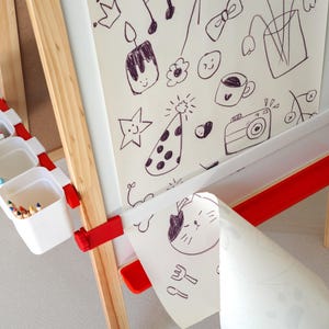 May include: Wooden easel with a white drawing surface and red accents. The easel has a built-in tray with three white containers filled with art supplies. The drawing surface features black marker doodles, including a star, a camera, and a cat.