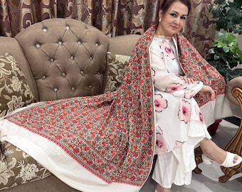 May include: A woman wearing a white floral print outfit and a red, white, and green floral shawl. The shawl is draped over the shoulders and extends to the sofa. The outfit includes a top, pants, and white heels.