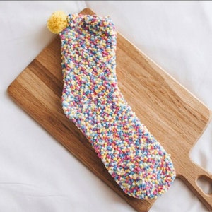 Peut inclure: Une paire de chaussettes moelleuses avec un motif de confettis colorés et un pompon jaune sur le dessus.