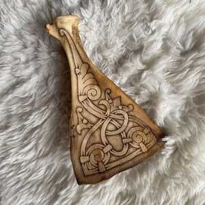 May include: A carved bone artifact with intricate Celtic knotwork and swirling patterns. The object has a light brown color with darker etched details. The piece is displayed on a white, fluffy surface.